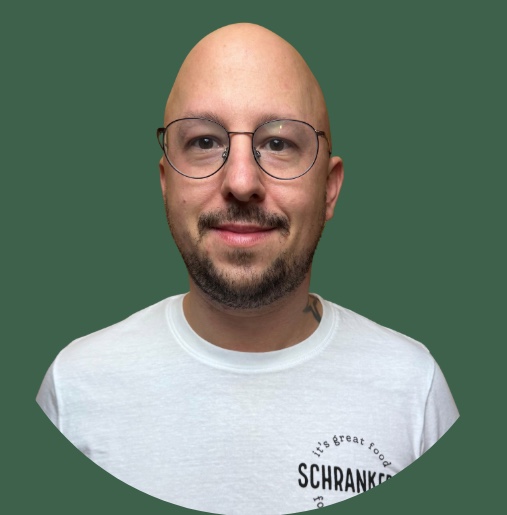 Lächelnder Mann mit Brille im Schrankerl T-Shirt für moderne Mitarbeiterverpflegung vor grünem Hintergrund.