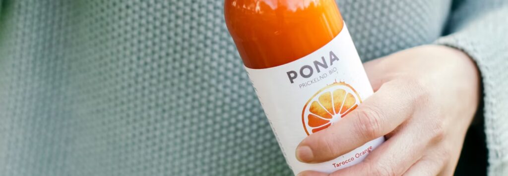 Eine Hand hält eine Flasche Pona Tarocco Orange für die Schrankerl Mitarbeiterverpflegung.