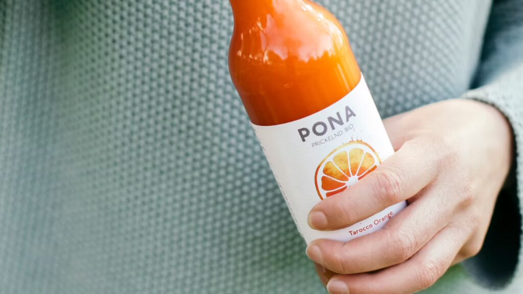 Eine Hand hält eine Flasche Pona Tarocco Orange als gesunde Mitarbeiterverpflegung.