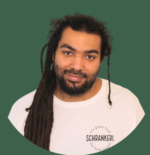 Lächelnder Mitarbeiter von Schrankerl mit Dreadlocks im weißen T-Shirt vor grünem Hintergrund.