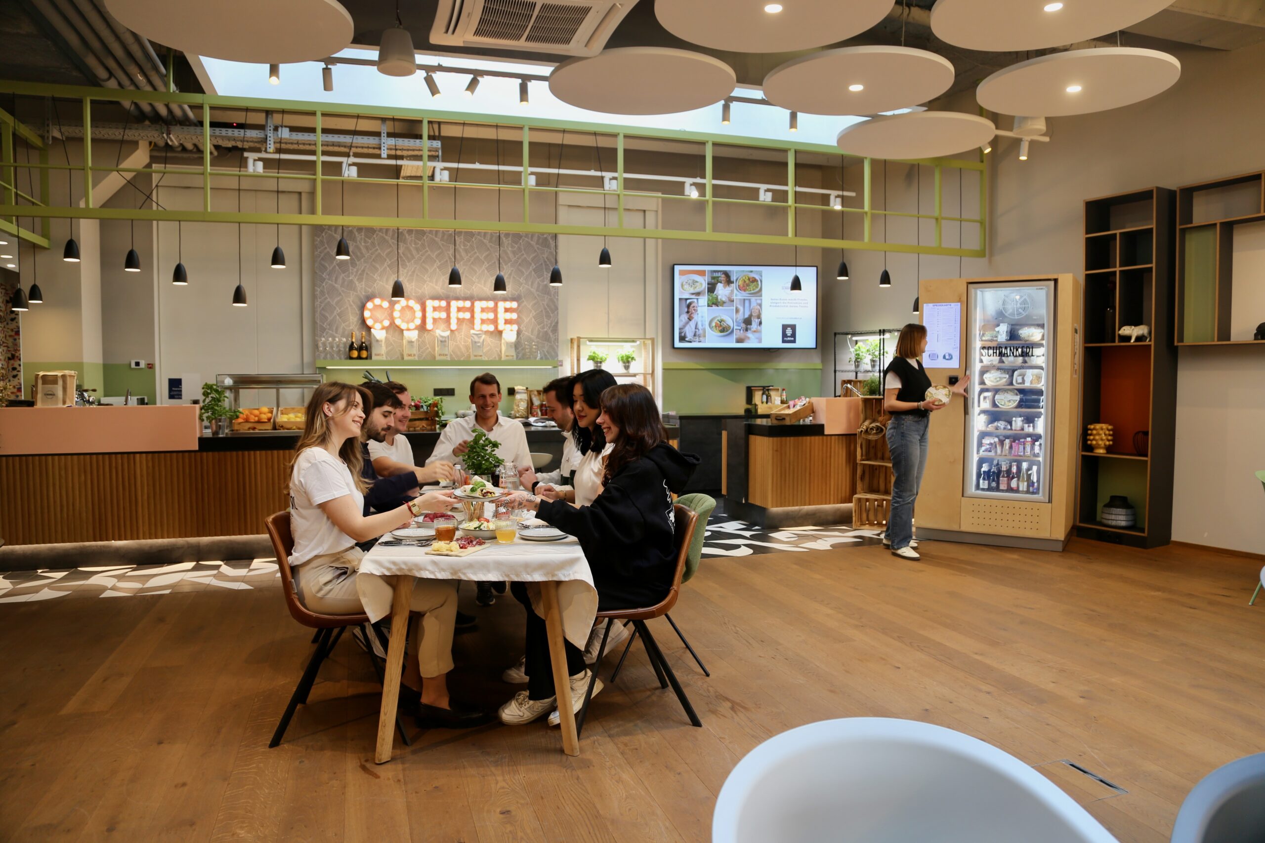 Employees enjoying fresh employee catering together at a dining table in a modern break room with a Schrankerl refrigerator.